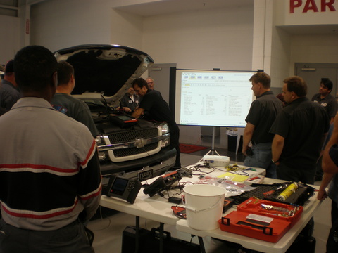 Technicians around a truck with its hood up, scan data on a screen and tools on the bench