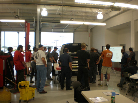 A class standing around a truck with its hood up in a bright shop