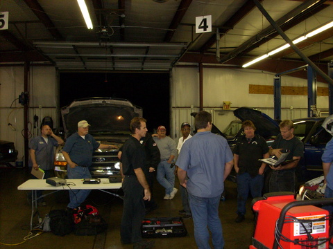 A hands-on class gathered around a truck with its hood up in a shop bay